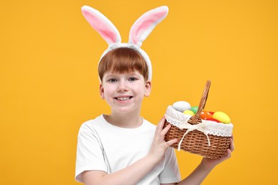 Photo of Easter celebration. Cute little boy with bunny ears and wicker basket full of painted eggs on orange background