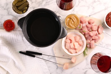 Photo of Fondue pot with oil, forks, raw meat pieces, glasses of red wine and other products on white marble table, flat lay