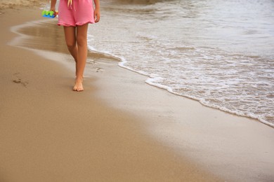 Photo of Little girl with plastic toys walking on sandy beach near sea, closeup. Space for text