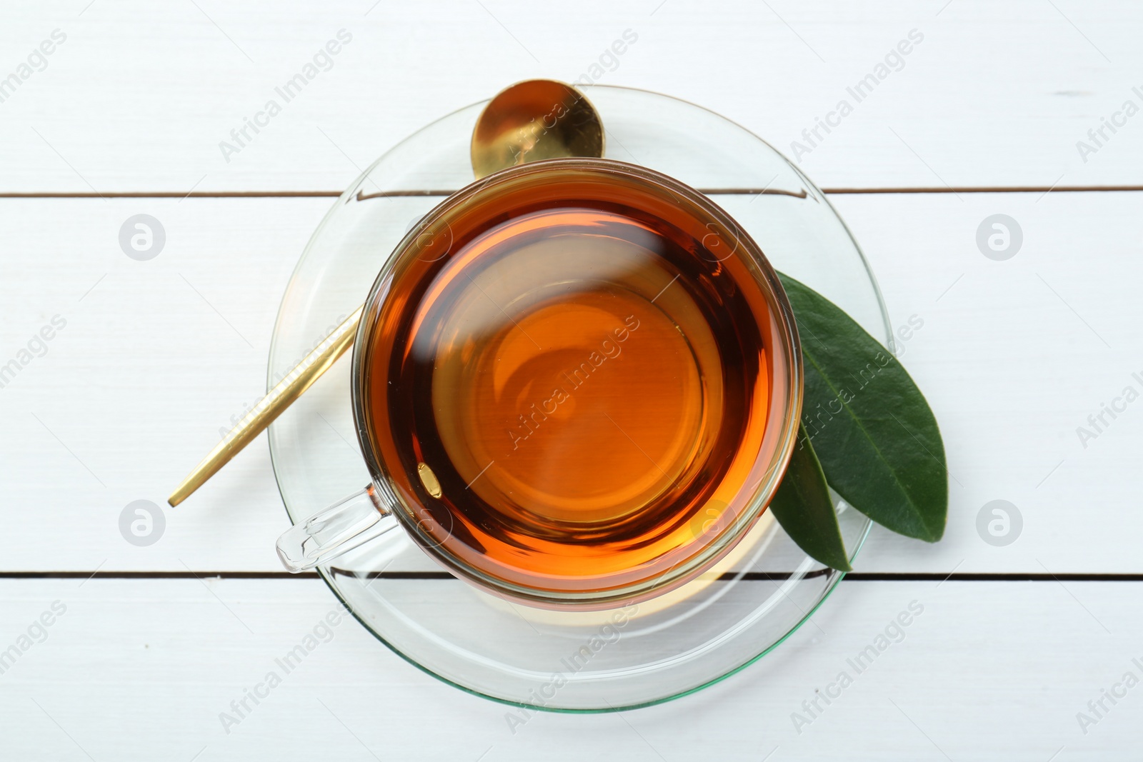 Photo of Aromatic tea in glass cup, spoon and green leaves on white wooden table, top view