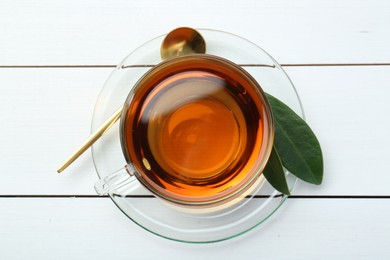 Aromatic tea in glass cup, spoon and green leaves on white wooden table, top view