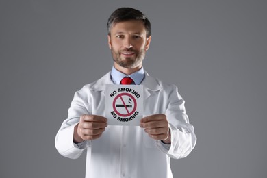 Doctor holding card with no smoking sign on gray background, selective focus