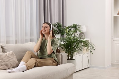 Beautiful young woman with headphones listening music on sofa in room with green houseplants