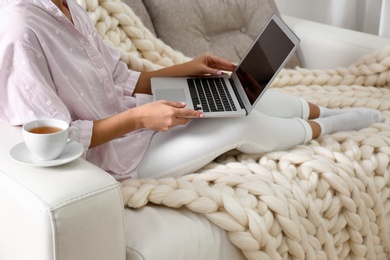 Photo of Woman using laptop on couch with soft knitted blanket at home, closeup