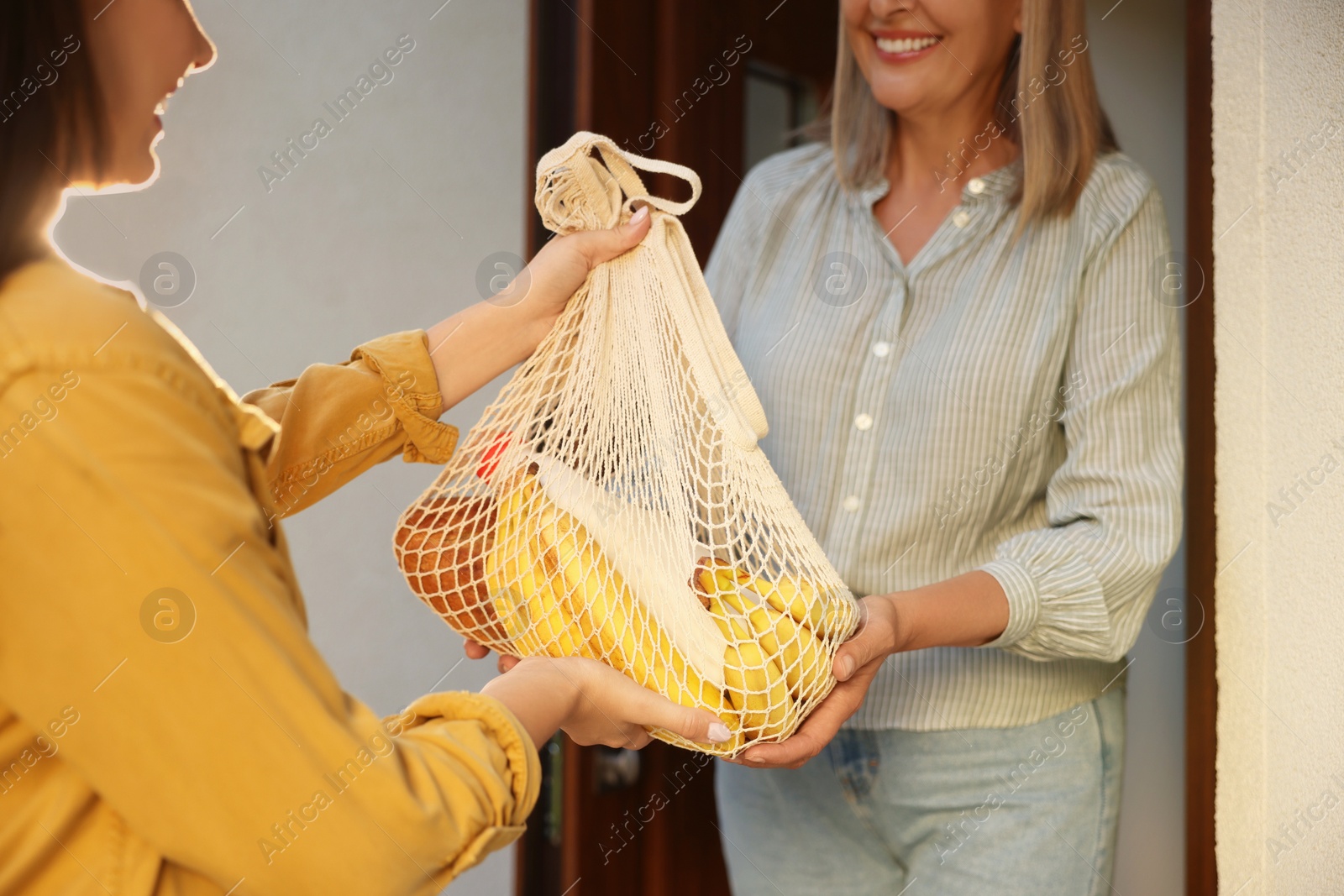 Photo of Young woman with net bag of products helping her senior neighbour outdoors on sunny day, closeup