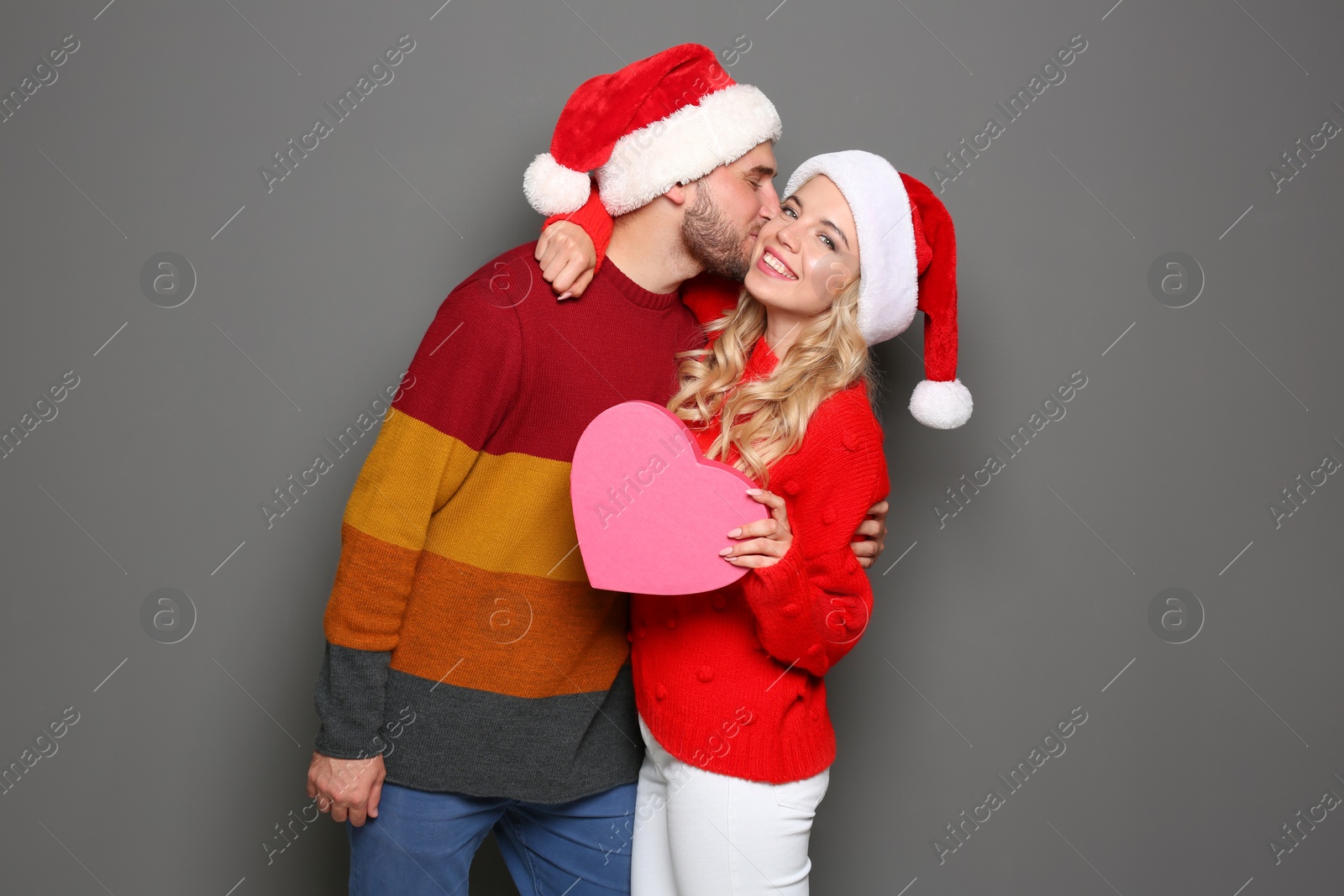 Photo of Young couple in Santa hats with pink heart on grey background. Christmas celebration