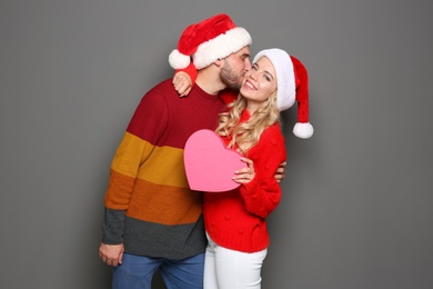 Young couple in Santa hats with pink heart on grey background. Christmas celebration