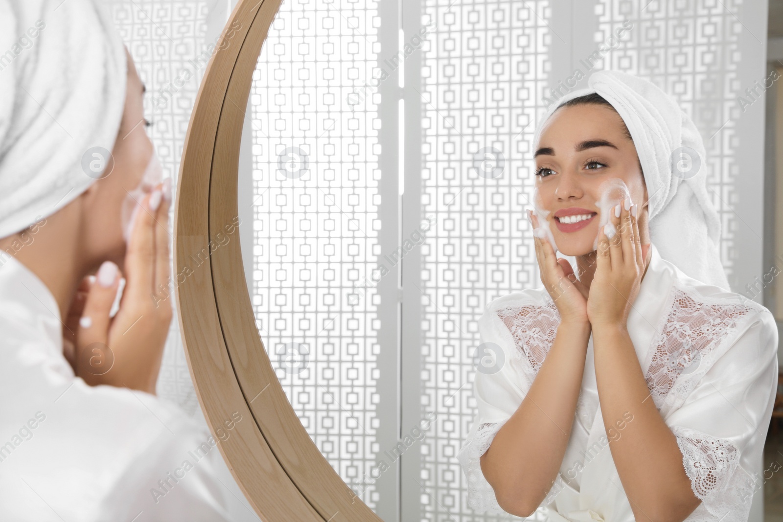 Photo of Beautiful young woman applying cleansing foam onto face near mirror in bathroom. Skin care cosmetic