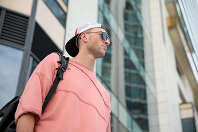 Handsome young man with stylish sunglasses and backpack near building outdoors, low angle view. Space for text