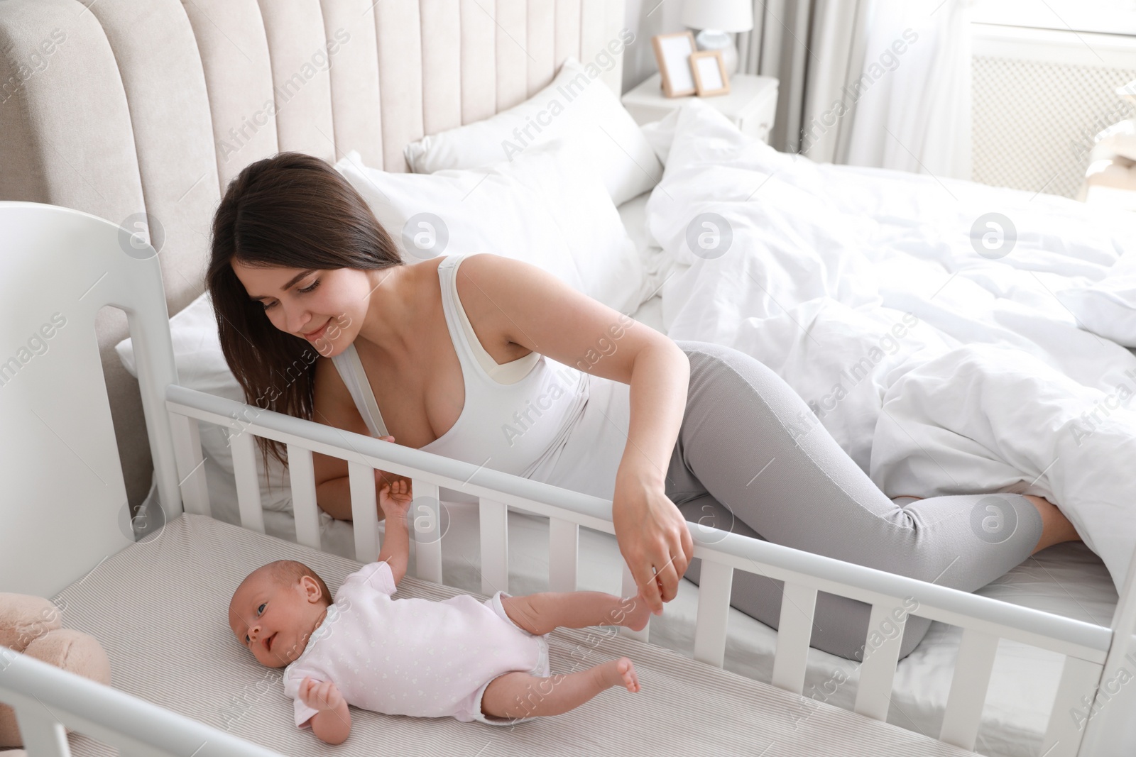 Photo of Happy young mother near bedside crib with her cute newborn baby at home