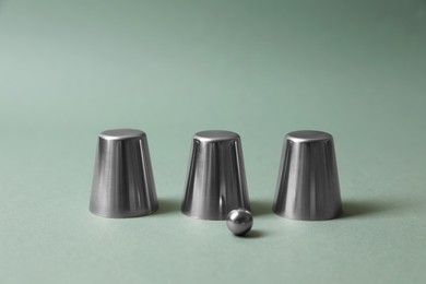 Three metal cups and ball on pale olive background. Thimblerig game