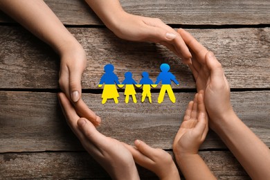 Image of Parents and child protecting paper family cutout in colors of Ukrainian flag at wooden table, top view