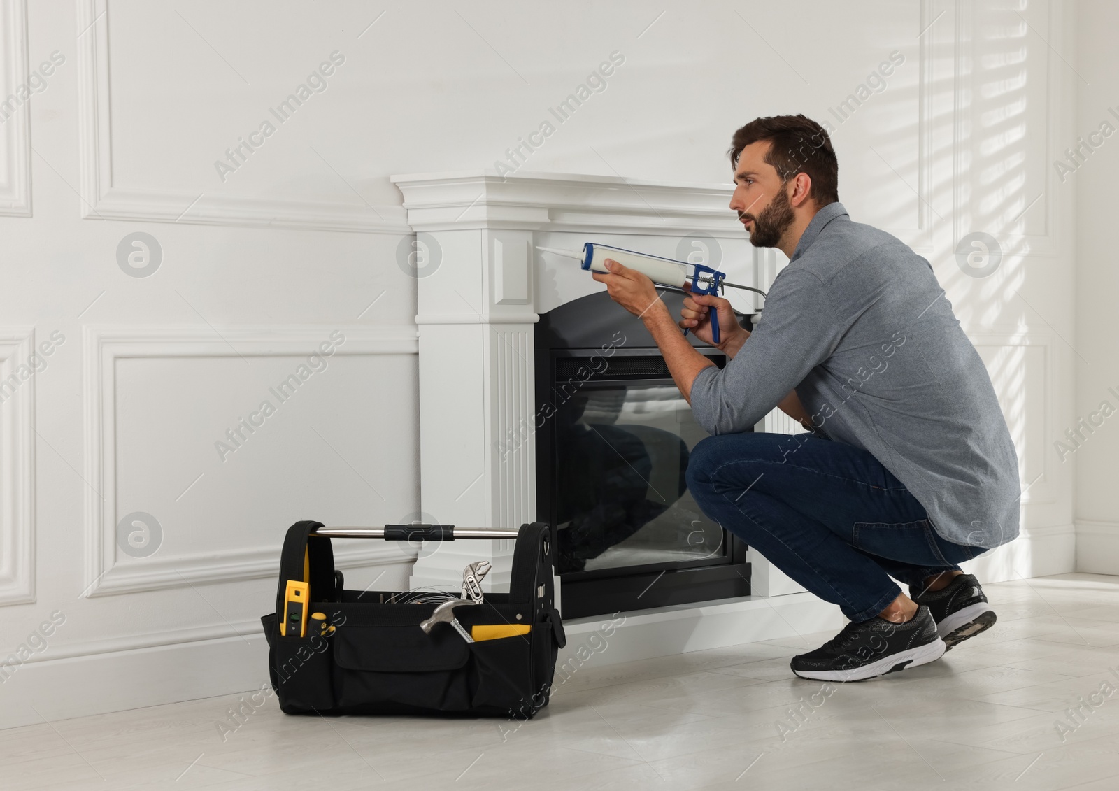 Photo of Man sealing electric fireplace with caulk near white wall in room