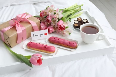 Photo of Tasty breakfast served in bed. Delicious desserts, tea, flowers, gift box and card with phrase I Love You on tray