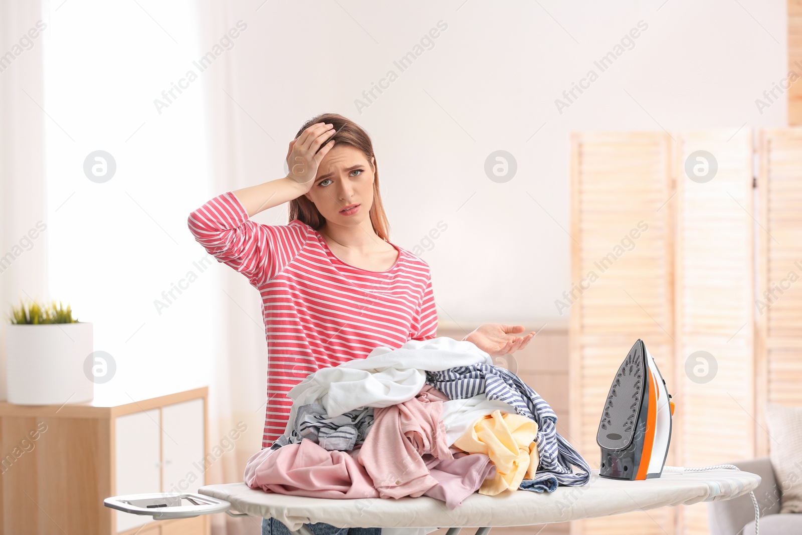 Photo of Young tired woman with clean laundry at ironing board indoors