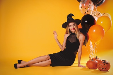 Beautiful woman in witch costume with balloons and pumpkins on yellow background, space for text. Halloween party