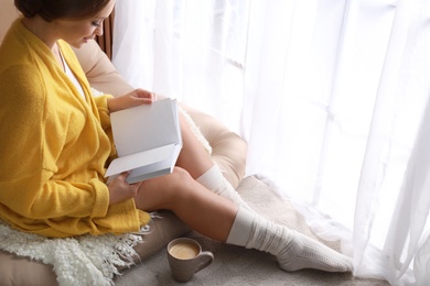 Beautiful young woman with cup of coffee reading book near window at home, space for text. Winter atmosphere