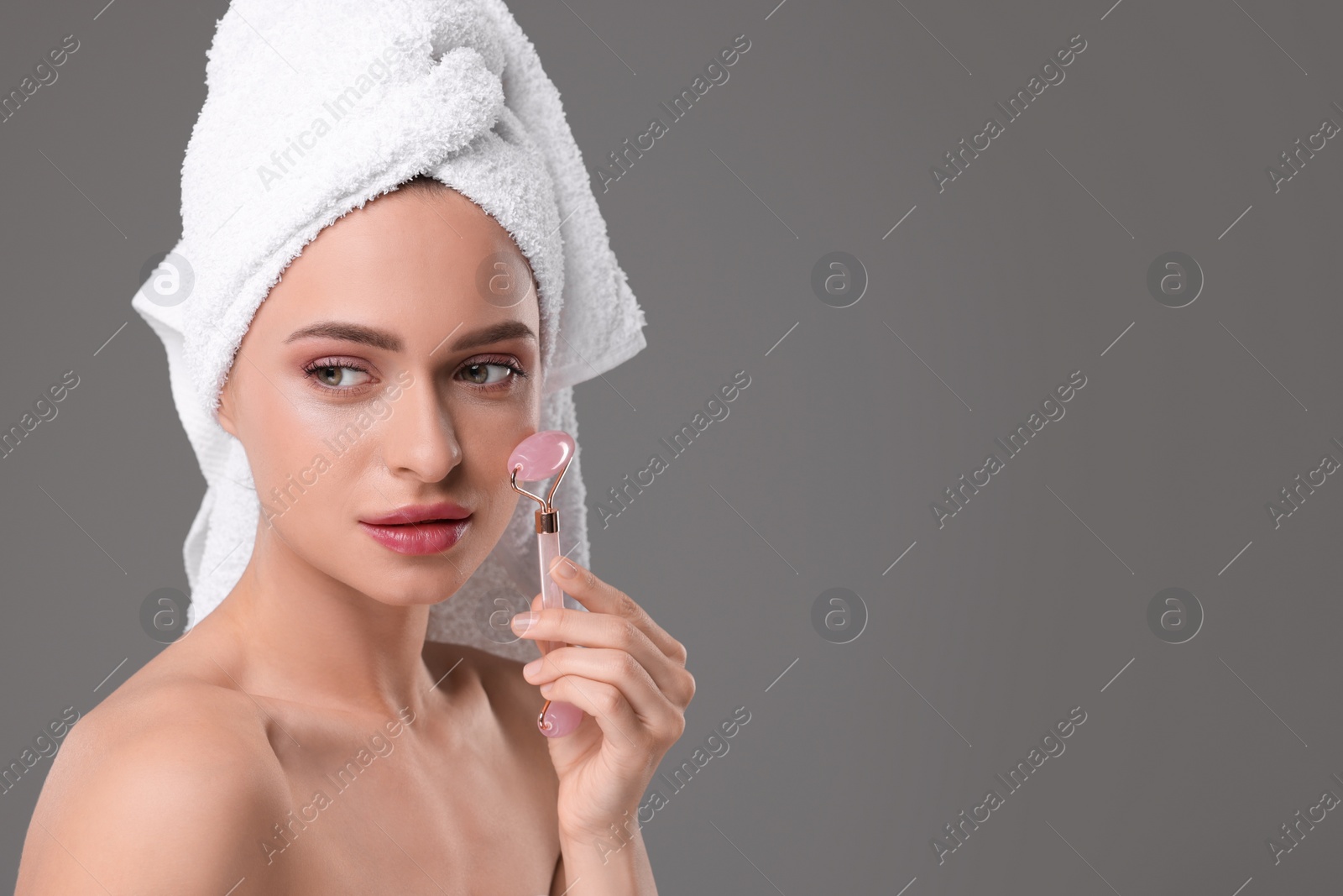 Photo of Young woman massaging her face with rose quartz roller on grey background, space for text