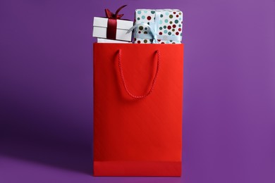 Red paper shopping bag full of gift boxes on purple background