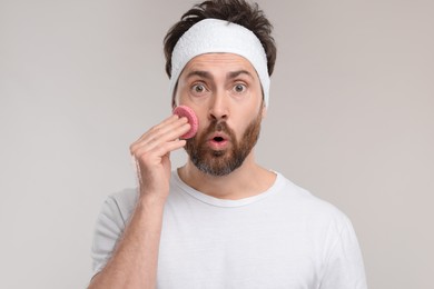 Emotional man with headband washing his face using sponge on light grey background