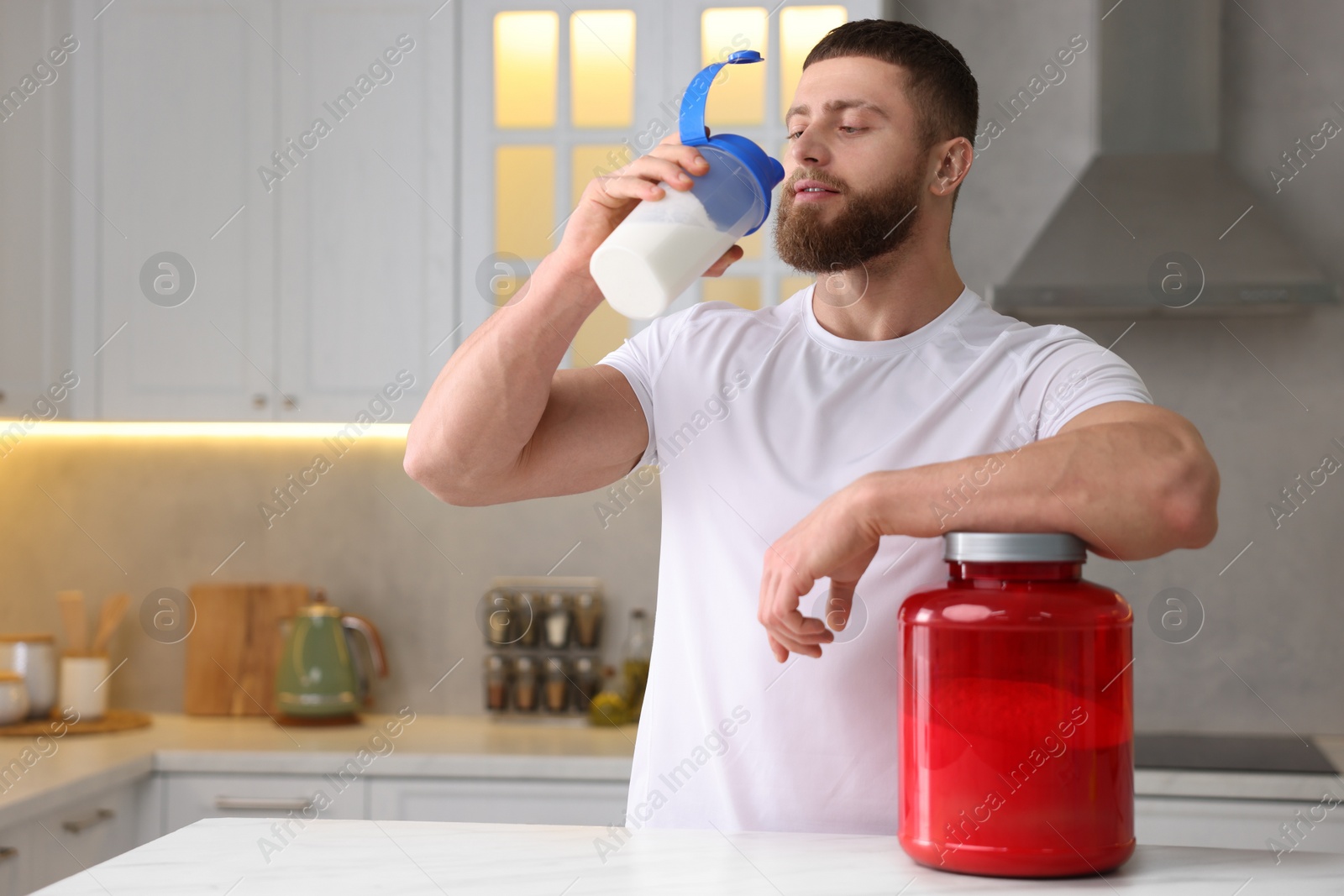 Photo of Young man with shaker of protein and powder at white marble table in kitchen