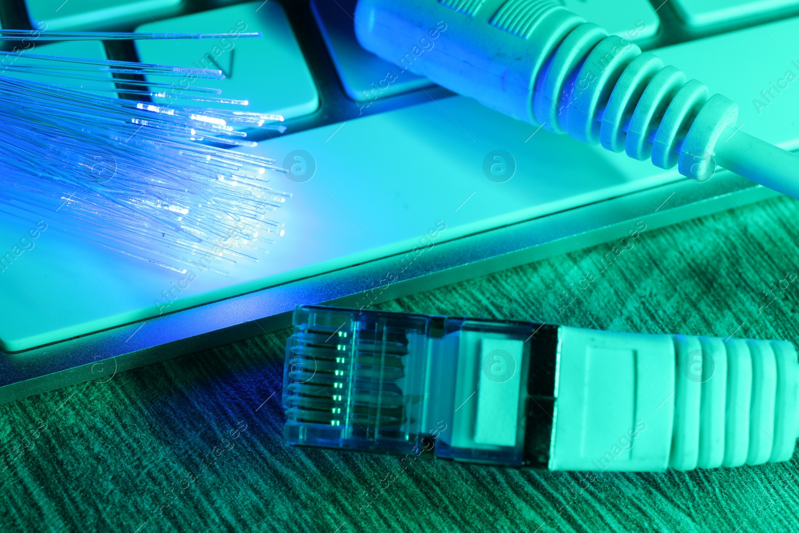 Photo of Optical fiber strands transmitting color light near cable with modular connector and computer keyboard on table, closeup