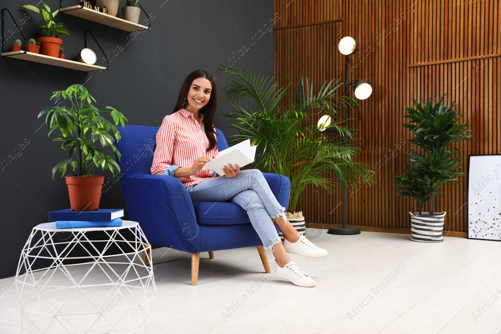 Photo of Happy woman reading book in armchair at home. Indoor plants for trendy interior design