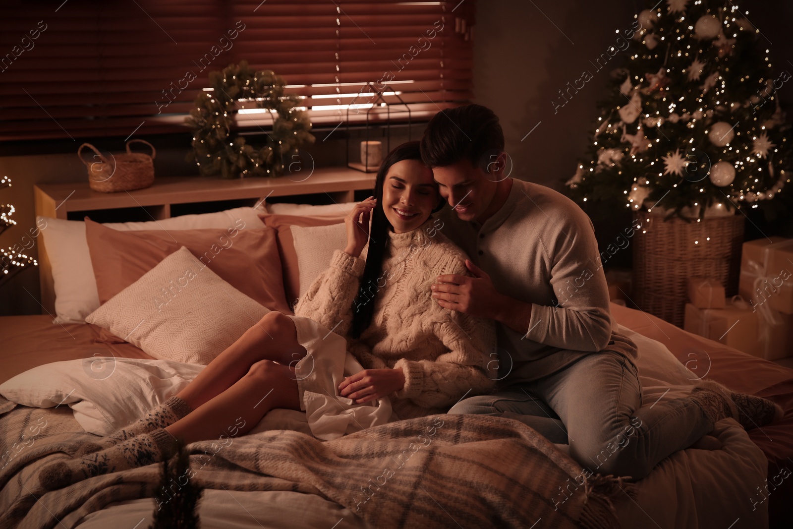 Photo of Happy couple sitting on bed in festively decorated room. Christmas celebration