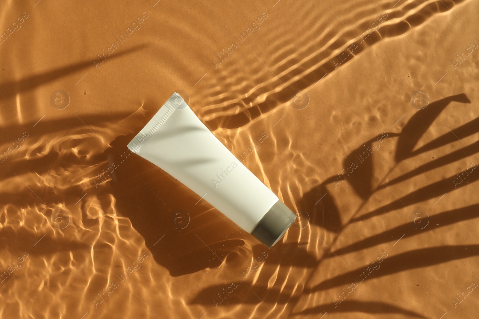 Photo of Tube with moisturizing cream in water and shadows from palm leaves on orange background, top view