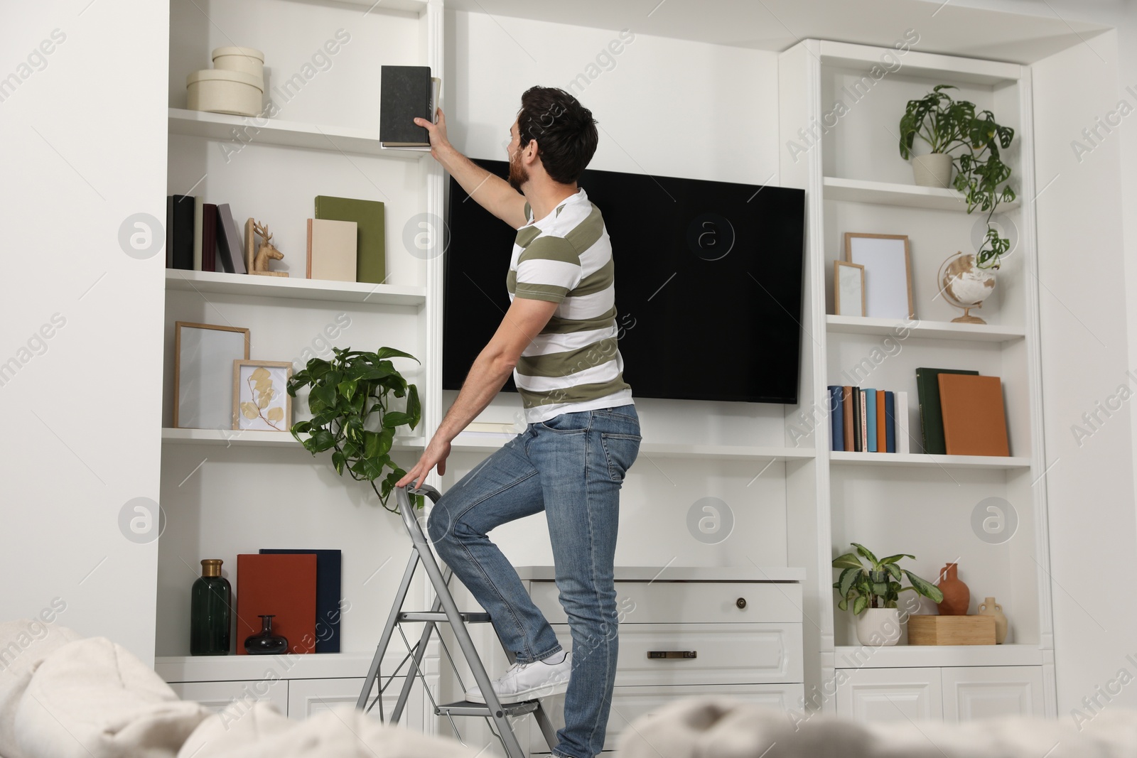 Photo of Man on metal folding ladder taking book from shelf at home