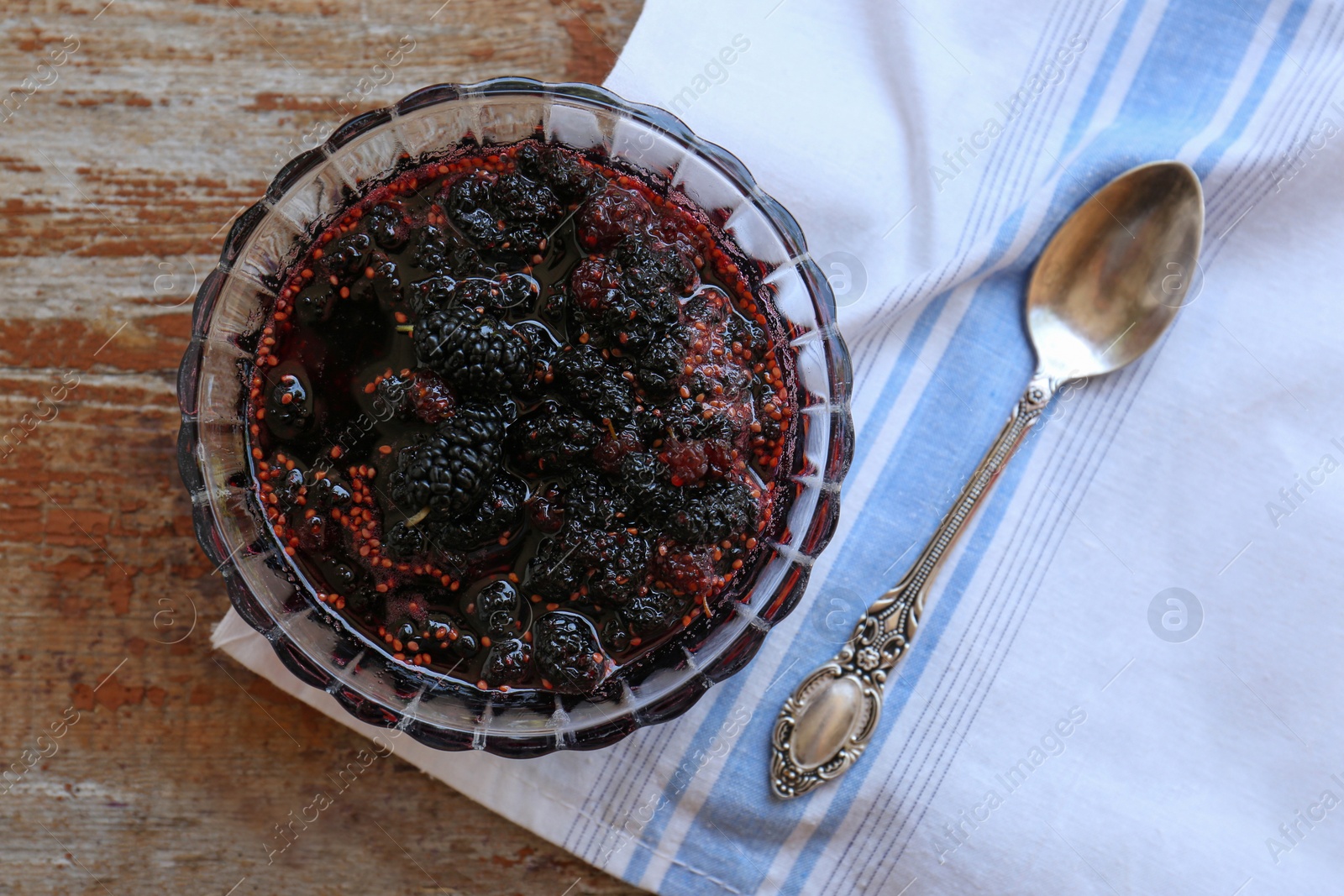 Photo of Bowl of sweet black mulberry jam, napkin and spoon on wooden table, top view