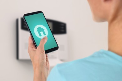 Image of Woman operating home alarm system via mobile phone against white wall with security control panel, closeup. Application with illustration of house on device screen