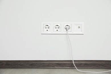 Power sockets with inserted plug on white wall indoors. Electrical supply