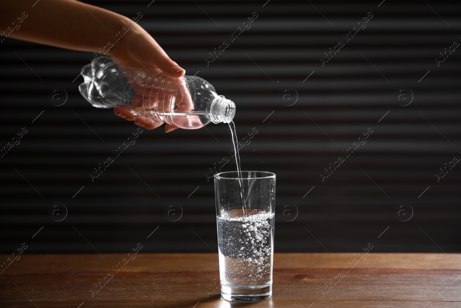 Photo of Woman pouring water from bottle into glass on wooden table against dark background, closeup