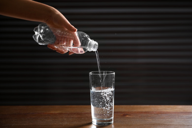 Photo of Woman pouring water from bottle into glass on wooden table against dark background, closeup