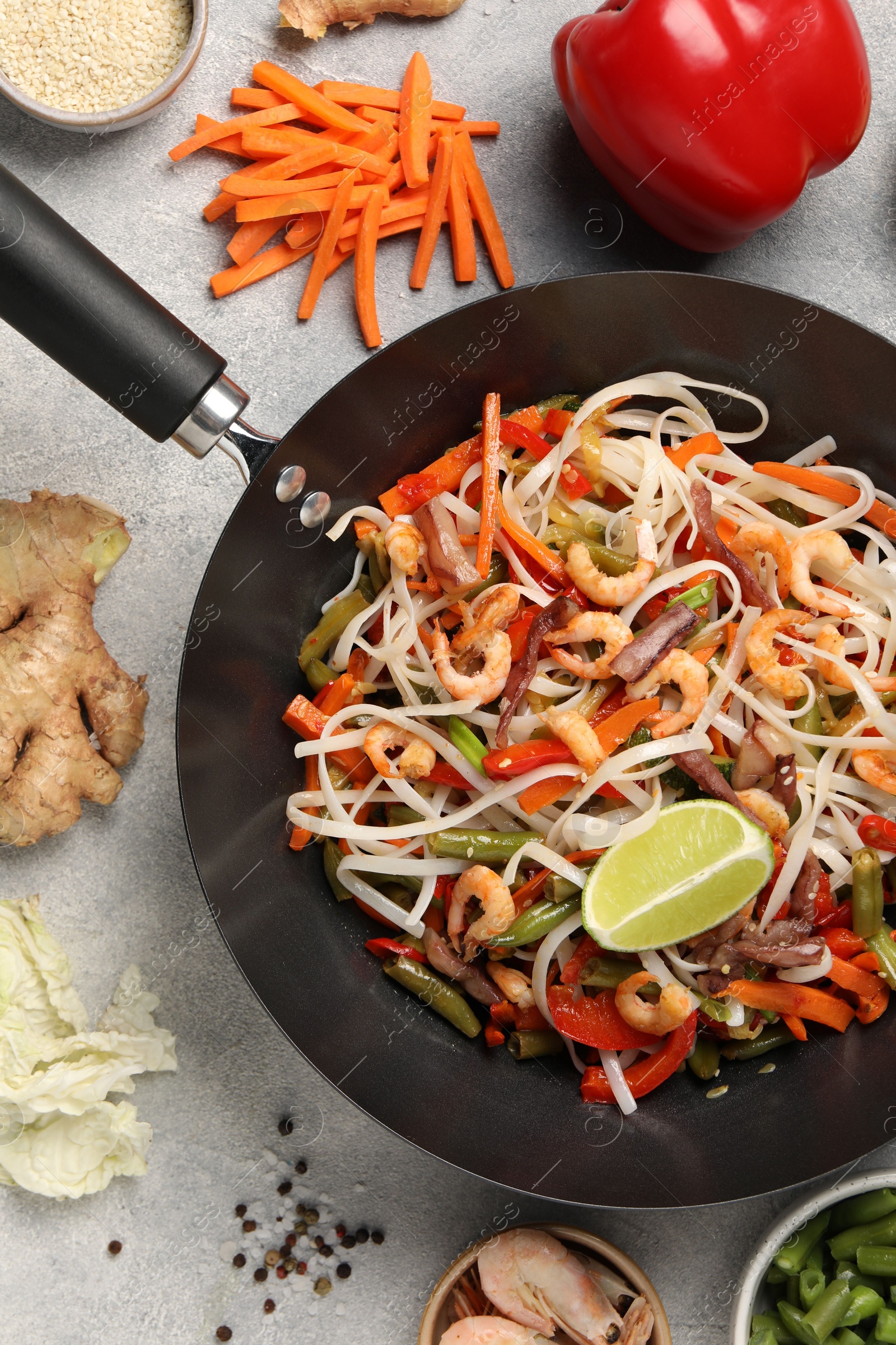 Photo of Shrimp stir fry with noodles and vegetables in wok surrounded by ingredients on grey table, flat lay