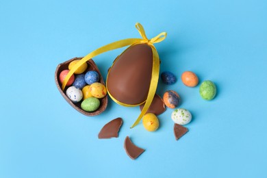 Tasty whole chocolate egg with yellow bow and different candies on light blue background, above view