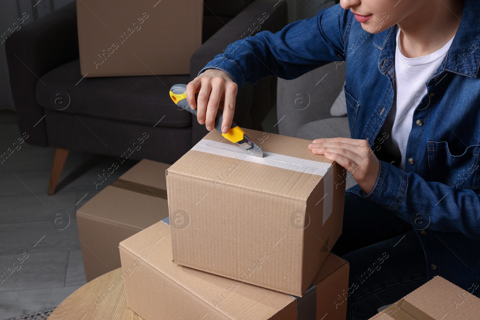 Photo of Young woman using utility knife to open parcel at home, closeup