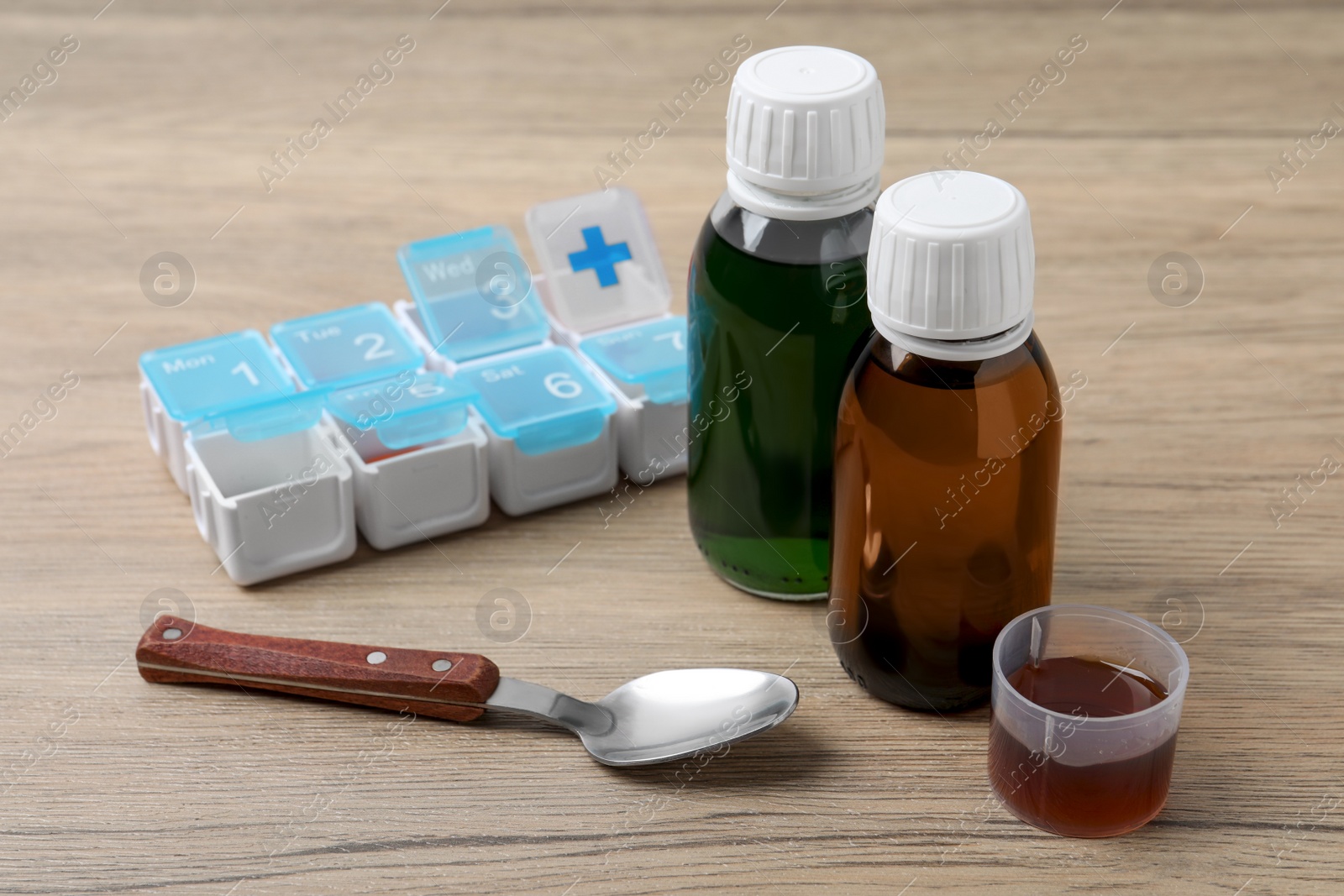 Photo of Bottles of syrup, measuring cup, spoon and weekly pill organizer on wooden table. Cold medicine
