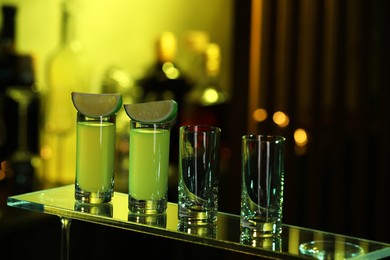 Shot glasses with alcohol drink and lime wedges on mirror surface against blurred background