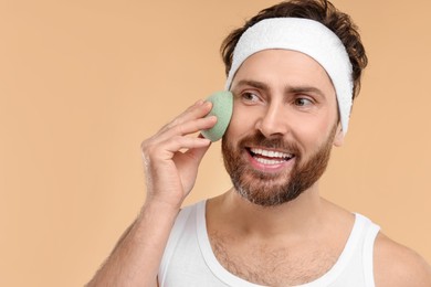 Man with headband washing his face using sponge on beige background, space for text