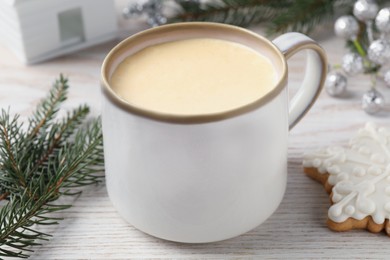 Cup of delicious eggnog, fir branch and cookie on wooden table, closeup