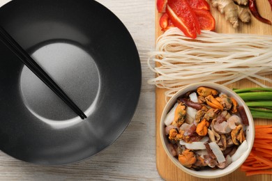 Photo of Black wok, chopsticks and board with products on light wooden table, top view