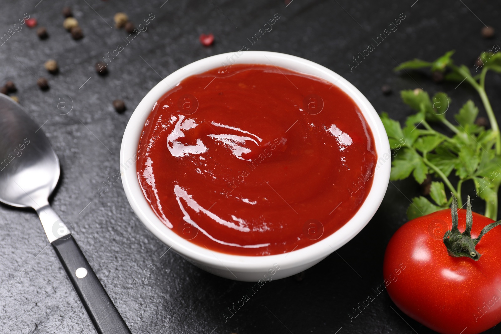 Photo of Organic ketchup in bowl, fresh tomato and spoon on black table, closeup. Tomato sauce