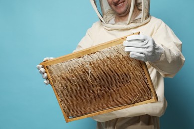 Beekeeper in uniform holding hive frame with honeycomb on light blue background, closeup