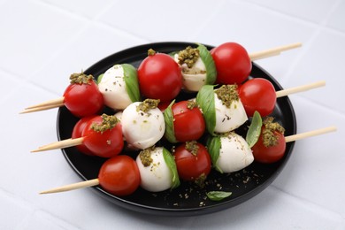 Photo of Caprese skewers with tomatoes, mozzarella balls, basil and pesto sauce on white tiled table, closeup