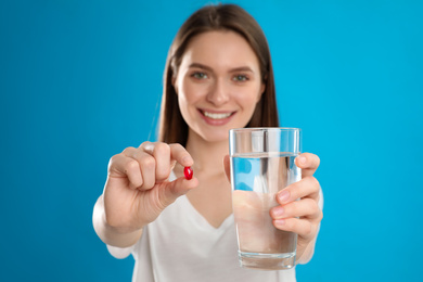 Young woman with vitamin pill and glass of water against blue background, focus on hands