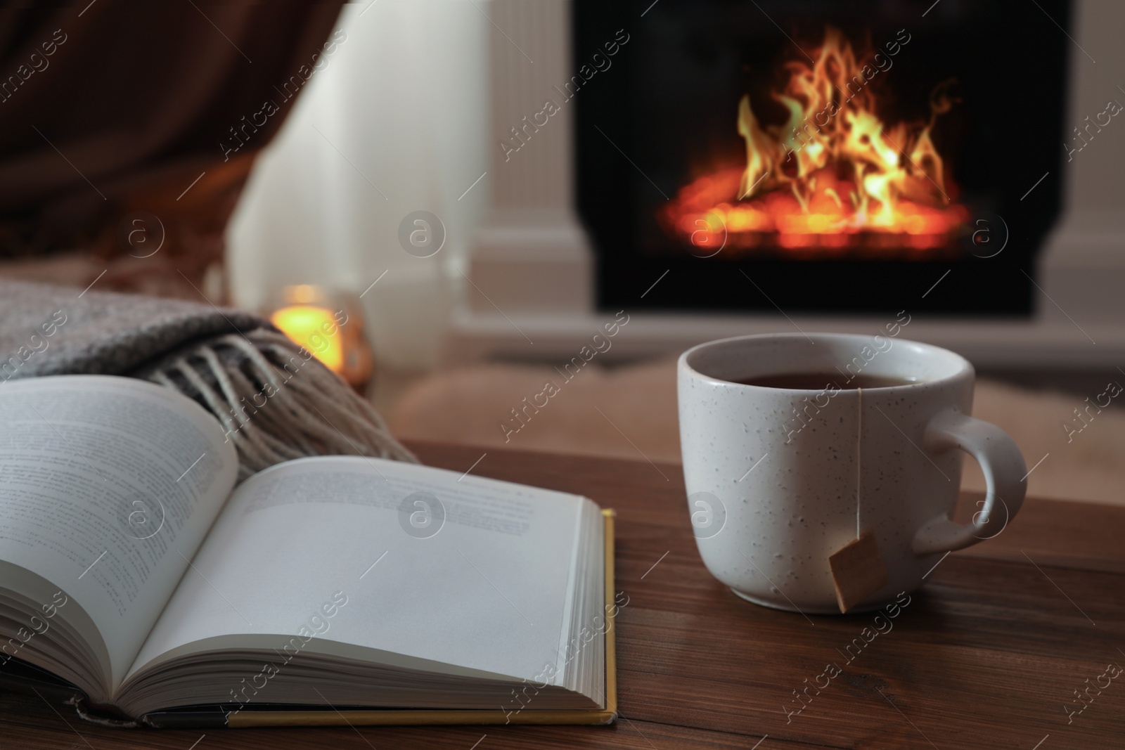 Photo of Cup of hot tea and book on wooden table near fireplace at home. Cozy atmosphere