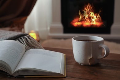 Cup of hot tea and book on wooden table near fireplace at home. Cozy atmosphere