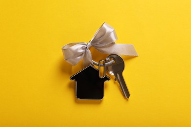 Key with trinket in shape of house and grey bow on yellow background, top view. Housewarming party